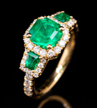 Yellow Gold 3 Stone Emerald Ring on a black background.