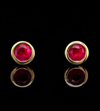 Round Red Beryl Bezel Set Studs in yellow gold front view on a black reflective surface.